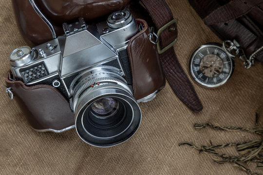 Top view of a vintage photo camera and a brown leather bag with scarf and pocket watch on sack cloth background.
