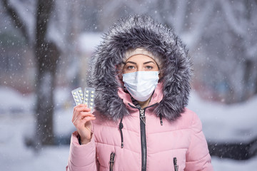 Fototapeta na wymiar Portrait of a beautiful girl in a pink jacket in a medical protective mask in holding yellow pills in her hands for colds and flu. Winter street portrait of a woman under snowfall.