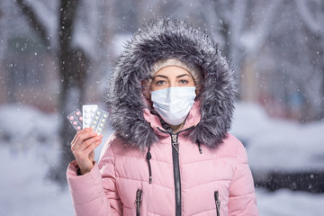 Fototapeta na wymiar Portrait of a beautiful girl in a pink jacket in a medical protective mask in holding white, pink, yellow pills in her hands for colds and flu. Winter street portrait of a woman under snowfall
