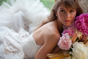 Bride in beautiful dress lying on sofa whit peonies in white studio interior. Romantic wedding style shot with sun light