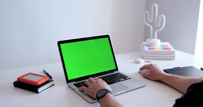 Young man working on a laptop with green screen chroma key in a white urban minimalist environment. Hands programming, pressing, clicking on a keyboard and using a mouse in a creative office workspace