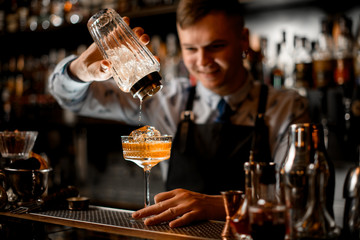 smiling young bartender pours finished cocktail from glassy shaker into glass with ice.