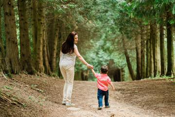 Fototapeta premium Backview of Mother walking with cute son in summer park. Happy woman with cheerful little boy outdoors