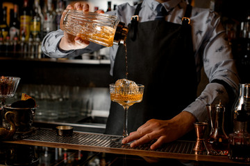 Professional bartender pours finished cocktail from glassy shaker into glass with ice.