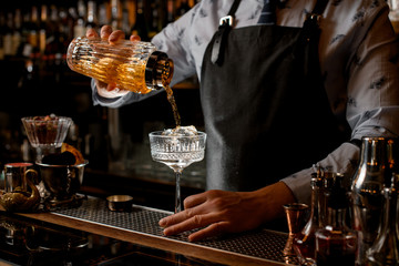 Professional barman pours finished cocktail from glassy shaker into glass with ice.