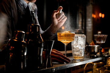 Professional bartender spray on cold brown alcoholic drink on bar counter.