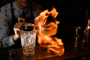 Close-up professional bartender spray and burn cold brown alcoholic drink on bar counter.