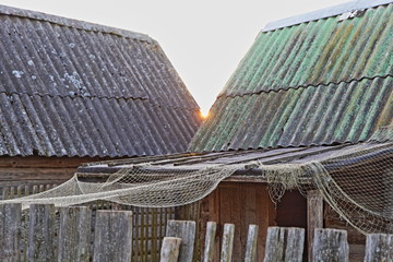 Old rustic view, sun between two roofs of weathered rural sheds with a drying net on wooden fence, fishing village yard
