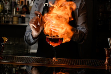 Bartender spray and burn cold brown alcoholic drink in wineglass.