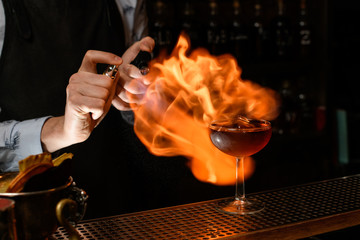 Bartender professionally spray and burn cold brown alcoholic drink in wineglass.