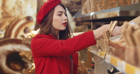 Fototapeta premium Beautiful Caucasian young woman in red hat and coat choosing and buying bakery. Charming stylish female customer buy bread in shop. Pretty girl buy baking.