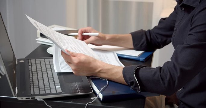 Close-up of male hands holding papers and making notes in the contract sitting at the table. Male entrepreneur reading documents, analyzing financial papers, preparing audit report at workplace