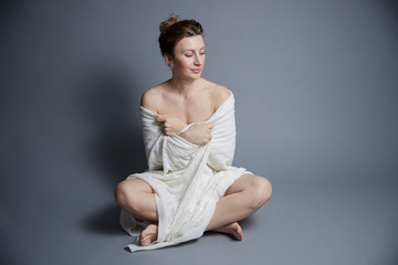 Portrait amazing slim cute beautiful young woman wrapped in white blanket with bare shoulders looks at camera on gray background