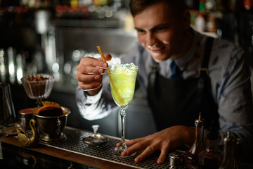 young smiling barman decorates by chopsticks wineglass with green cocktail.