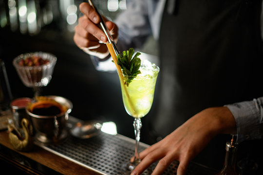 bartender carefully decorates by mint green alcoholic drink with ice in glass.