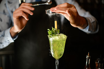 Close-up bartender decorates green alcoholic drink with ice in glass by powder.