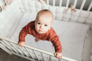 Beautiful little girl in a crib in a cozy light room in a sweater of fashionable shades. learns to stand at the support. cheerful little girl standing in the crib at home