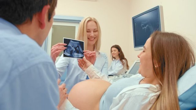 A young woman doctor shows a photo of an ultrasound scan of a child. A pregnant woman rests on a couch, a young couple happy and smiling