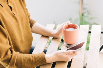 Close-up of woman hands holding ceramic cup of chamomi tea and is using mobile phone reading news, social network, doing online shopping while staying at home on quarantine period of corona virus 2020