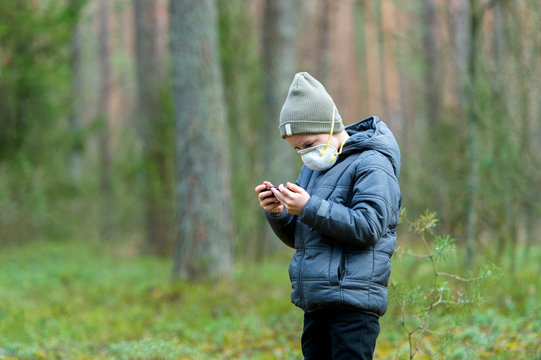 Kid With Mask In Their Mouths Due To Coronavirus Disease And Play Mobile Games On A Black Phone In A Forest