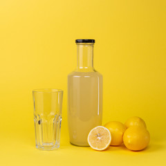 Bottle with lemon juice and a glass empty glass next to it and whole and sliced lemons isolated on a yellow background