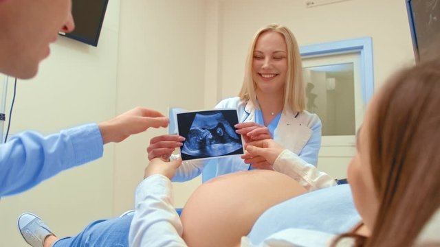 A young pregnant woman with her husband looks at her baby ultrasound. A doctor smiles at a happy couple