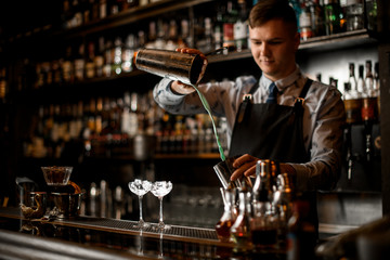 young handsome bartender pours cocktail in shaker. Two glasses with ice stand on bar.