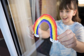 Girl Putting Picture Of Rainbow In Window At Home During Coronavirus Pandemic To Entertain Children