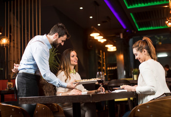 Smiling young female friends at a restaurant with waiter serving dinner