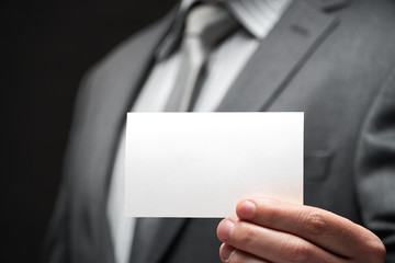 white blank business card closeup in businessman hand, gray suit, dark wall background