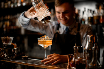 young smiling bartender pours drink from shaker into glass.