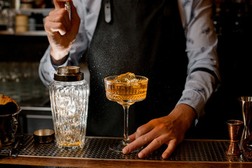 Barman in black apron carefully sprinkles on glass with brown alcoholic cocktail.