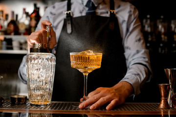 Bartender in black apron carefully sprinkles on glass with brown alcoholic cocktail.