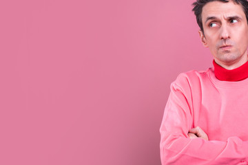 Cut view of man in pink sweater stand on right side and lok to left with crossed hands. Guy doubt with concentration. Isolated over pink background.