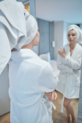 Fototapeta premium I am beautiful. Portrait of confident young woman looking at mirror with satisfaction while standing in bathrobe at home. Body care concept