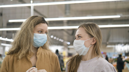 Two female friends put on medical masks in rubber gloves in a supermarket, sigh sadly and look at each other. Protective measures to combat the coronavirus pandemic.
