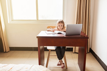 Pretty schoolgirl studying homework during her online lesson at home, social distance during quarantine covid-19, self-isolation, online education concept, home schooler. Workplace in living room.