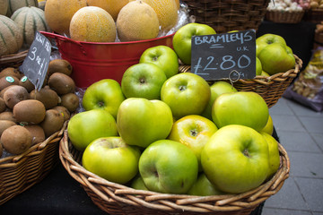 Green apples in wicker basket at a market for sale. In the background are counters with seasonal vegetables.