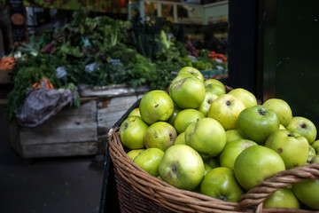 Green apples in wicker basket at a market for sale. In the background are counters with seasonal vegetables.