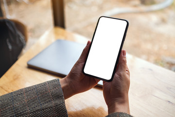 Mockup image of a woman holding mobile phone with blank white desktop screen with laptop on table