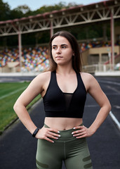 Young brunette woman, wearing black top and green leggings, standing on city stadium on summer morning, resting relaxing after training. Three-quarter portrait of sporty girl. Healthy life concept