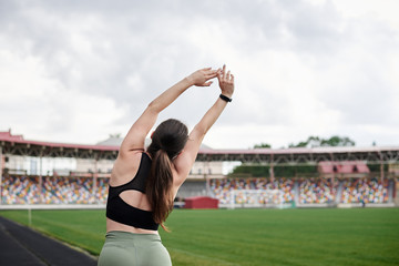 Young brunette sportswoman, wearing black top and green leggings, doing yoga pose on city stadium on summer morning, Three-quarter portrait of girl, stretching outside. Healthy active life concept.