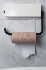 Finished roll of toilet paper with the inscription: you're screwed on a tiled bathroom