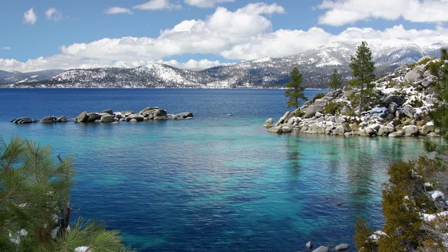 Lake Tahoe with snow on the ground and view on peaks of Sierra Nevada mountains, clear waters of alpine lake, clouds in the blue sky, 4k