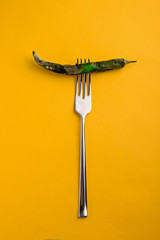 An ugly, rotten green chili pepper covered in mold is put on a metal fork on a yellow background with space for text in vertical orientation