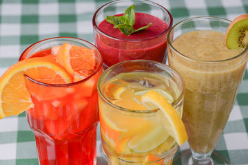 Four tropical mixed drinks, berries, orange and kiwi fruit cocktails served on green plaid tablecloth.