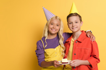 smiling kids with party caps holding plate with birthday cake and hugging on yellow background