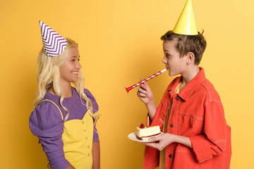 side view of boy blowing in party horn and giving plate with birthday cake to smiling friend with party cap on yellow background