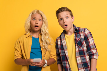 shocked kids holding smartphone and looking at camera on yellow background