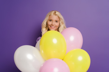 Fototapeta na wymiar smiling kid with balloons looking at camera on purple background